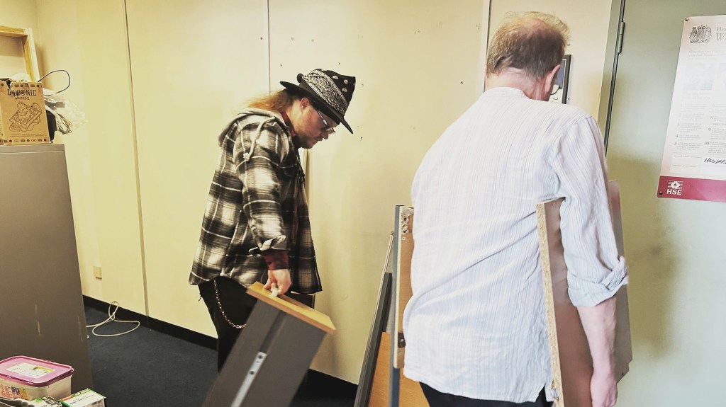 Lewins and Priestley are seen moving things into the new studio. Lewins is holding a filing cabinet drawer and Priestley is holding part of a desk. 