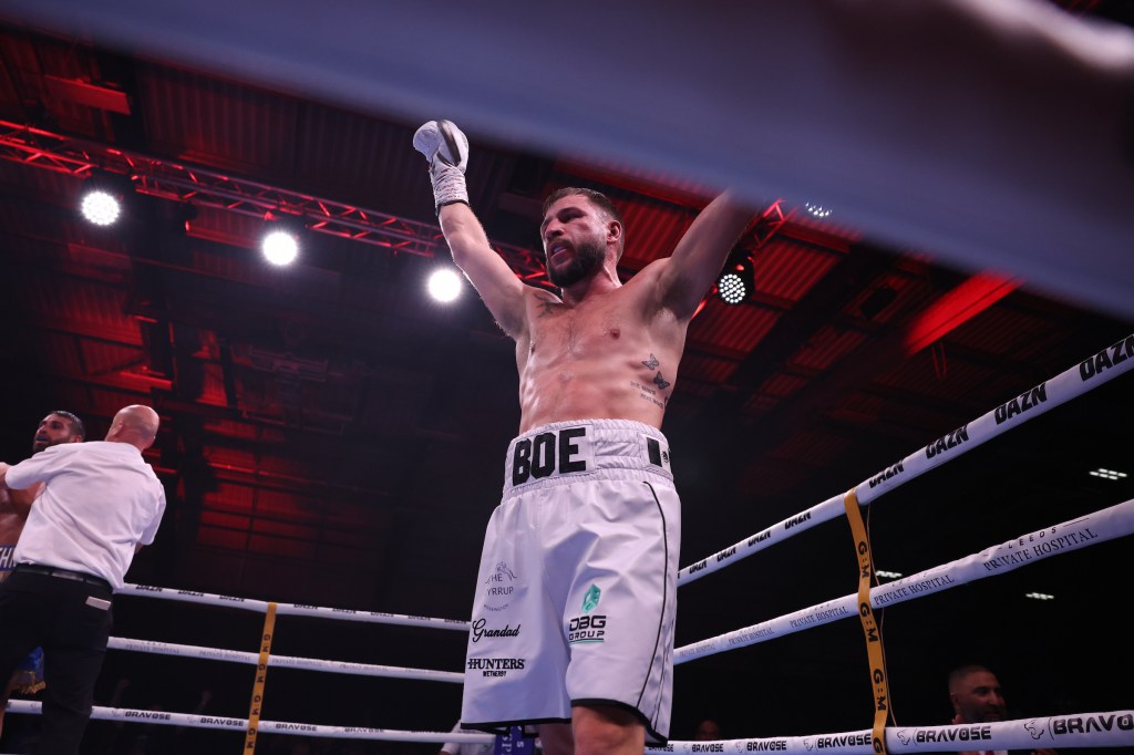 Maxi Hughes stands in the ring with his hands raised in celebration - he is sporting an all white attire.