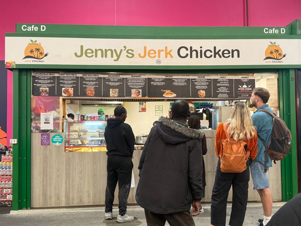 Jenny's Jerk Chicken food stall inside Kirkgate Market with a green painted front. Images of the menu items, including curry box meals, beef box meals, salad boxes, burgers, and wraps, are displayed around the opening hatch. Customers are visible near the stall, and staff members can be seen working in the kitchen behind the counter