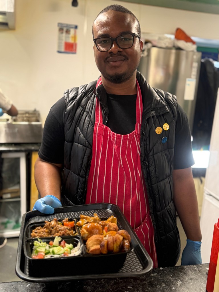A Jenny’s Jerk Chicken employee stands behind the counter smiling at the camera, holding a plate of dumplings, curry goat, and jerk chicken. The kitchen is visible in the background