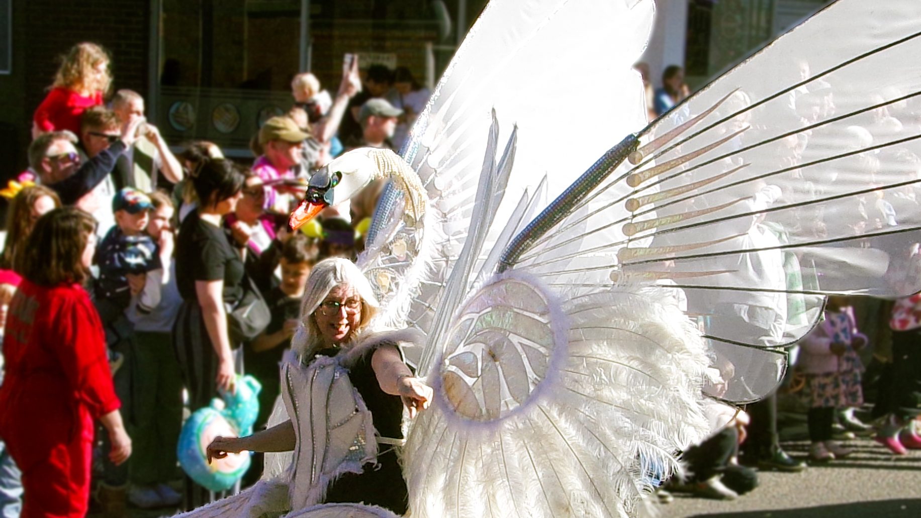 Alison Gardener, MP for Stoke-on-Trent South, wears a giant swan costume. 
The swan is attached to her back, and its head rises above her own. She is pointing to the crowd out of shot. She has white hair, which matches the swan's feathers.