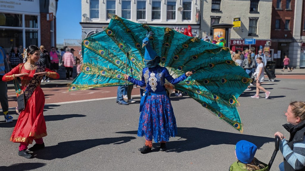 A drama group recreates the princess and the peacock fairytale. The princess wears a bright red flowing dress and black sash. 
The peacock wears a flowing blue and gold dress. Its head is worn as a hat by the performer, the performer holds out a long cape which represents the peacock's feathers.