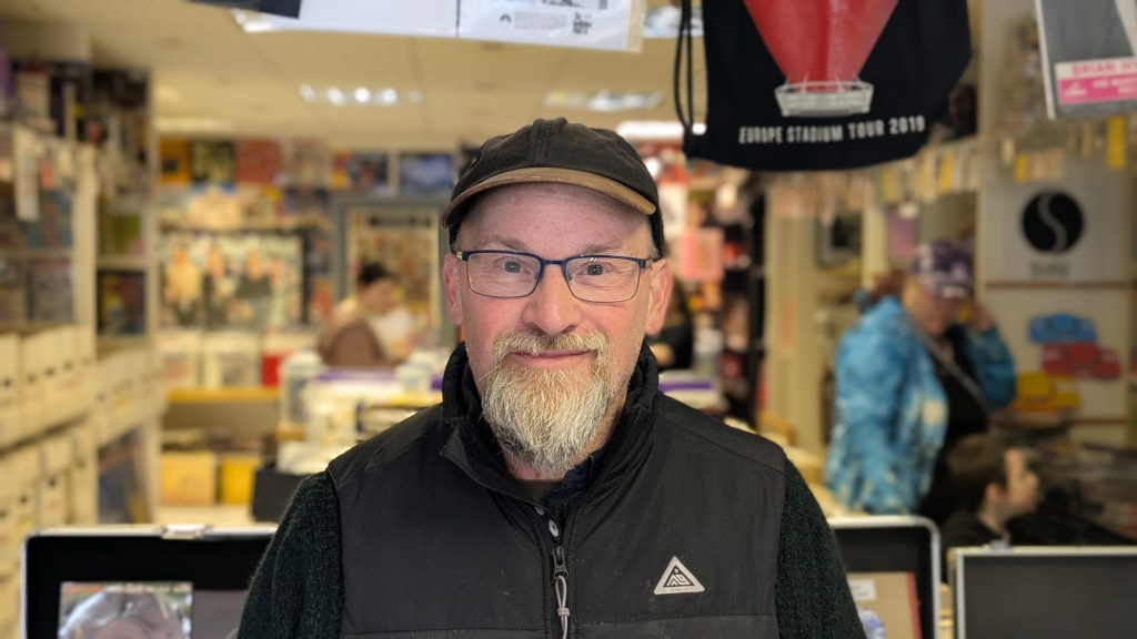 Dave Hawkins, the owner of Cannonball Records, poses in portrait. He wears a black cap with a black overcoat. He wears square black glasses and has a small white beard. 