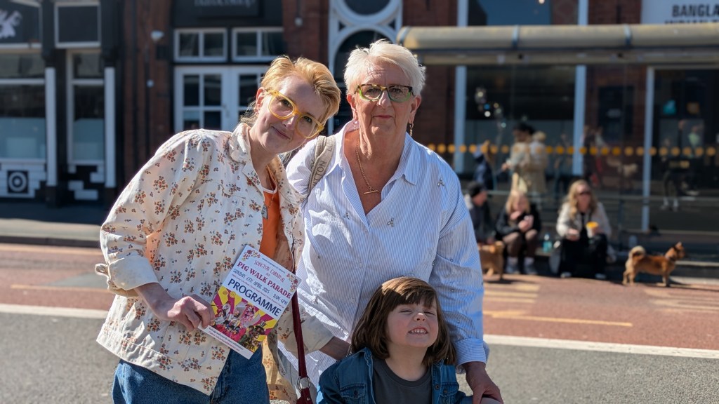 Charlotte, a young woman, poses with her family. 
Charlotte has short-swept over blonde hair and is wearing a flowery shirt with blue jeans with bright yellow glasses.
 Next to Charlotte is an older woman with the same hairstyle, with grey hair, who wears a white shirt and a big golden necklace. 
At the bottom of the picture poses a young boy with long brown hair, he grins to the camera.