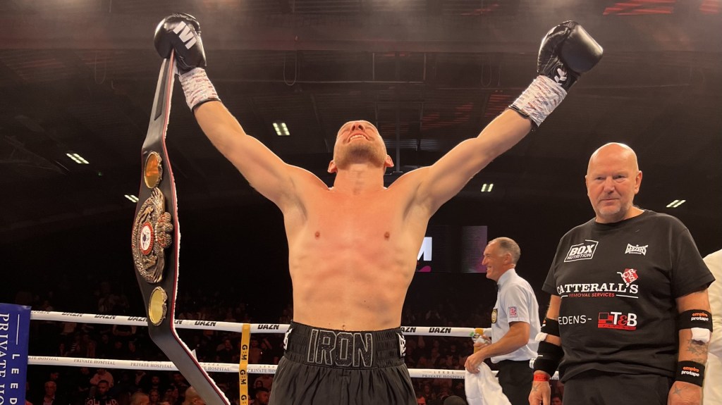 Ryan Walsh is stood with his belt and hands in the air as he looms up to the lights