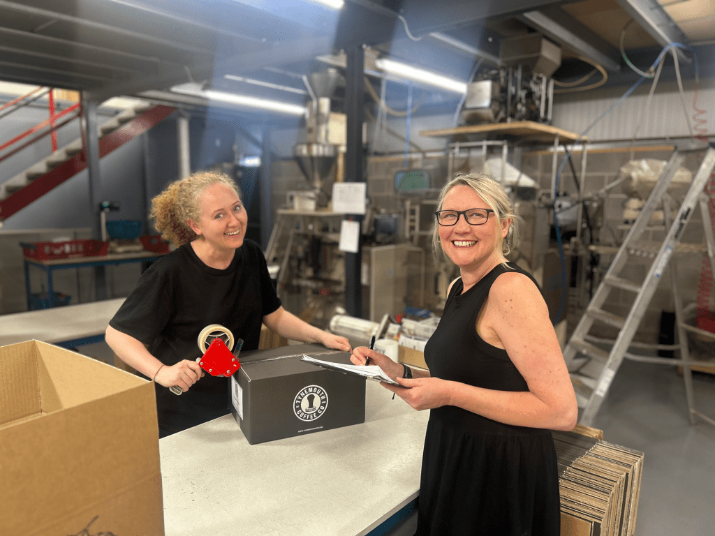 To women are stood over a table with a box of coffee between them. One of the women is taping the box closed while the other is writing on a clipboard