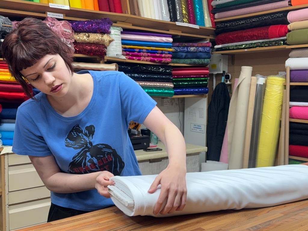 A woman folds a large piece of fabric at her market stall, surrounded by neatly stacked rolls of material behind her
