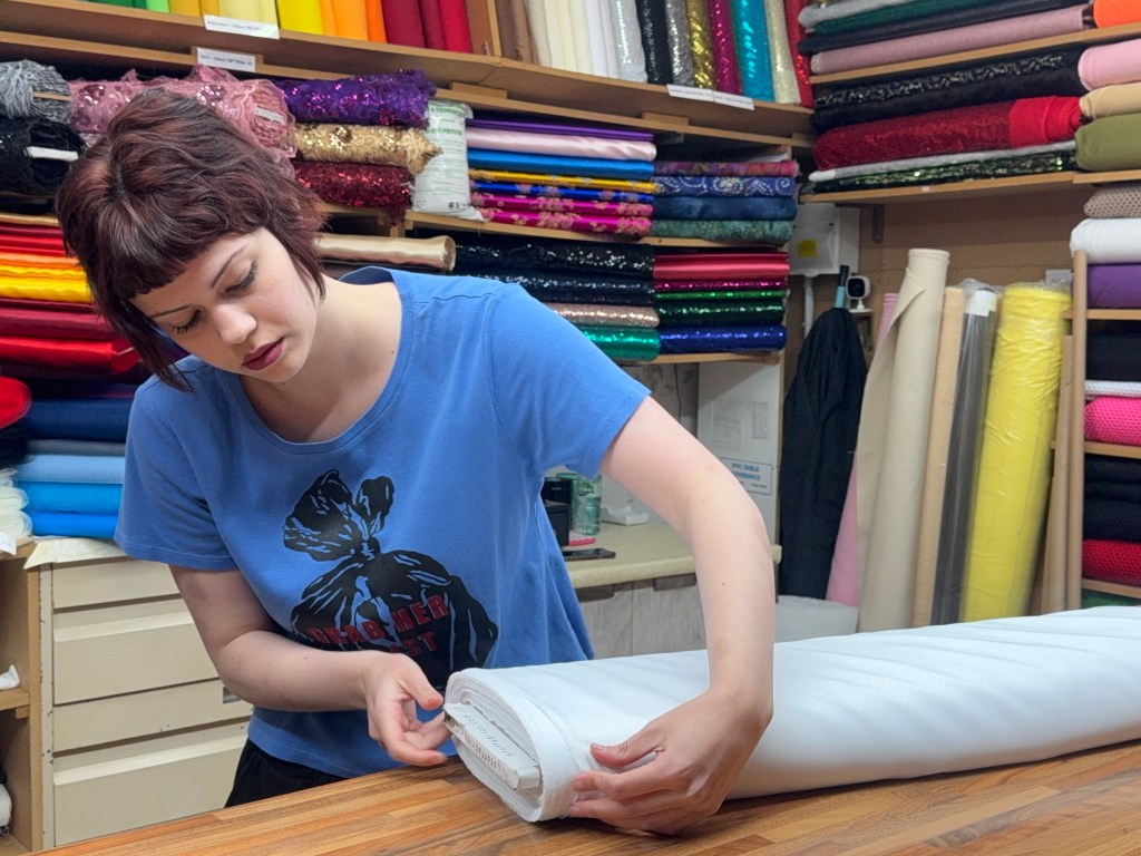 A woman folds a large piece of fabric at her market stall, surrounded by neatly stacked rolls of material behind her