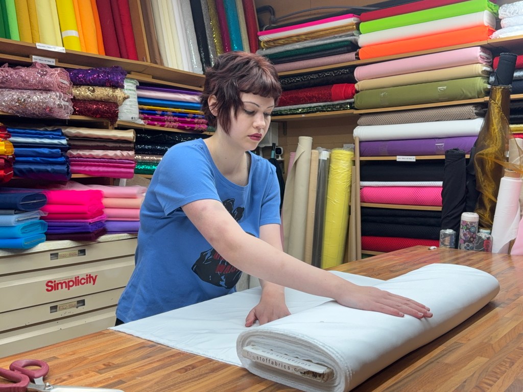 A woman folds a large piece of fabric at her market stall, surrounded by neatly stacked rolls of material behind her