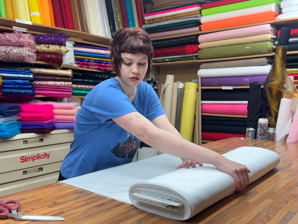 A woman folds a large piece of fabric at her market stall, surrounded by neatly stacked rolls of material behind her