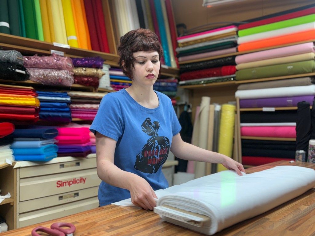 A woman folds a large piece of fabric at her market stall, surrounded by neatly stacked rolls of material behind her