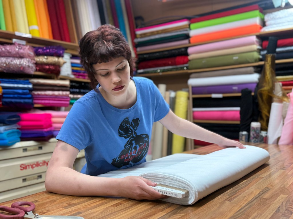 A woman folds a large piece of fabric at her market stall, surrounded by neatly stacked rolls of material behind her