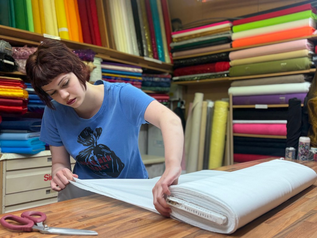 A woman folds a large piece of fabric at her market stall, surrounded by neatly stacked rolls of material behind her