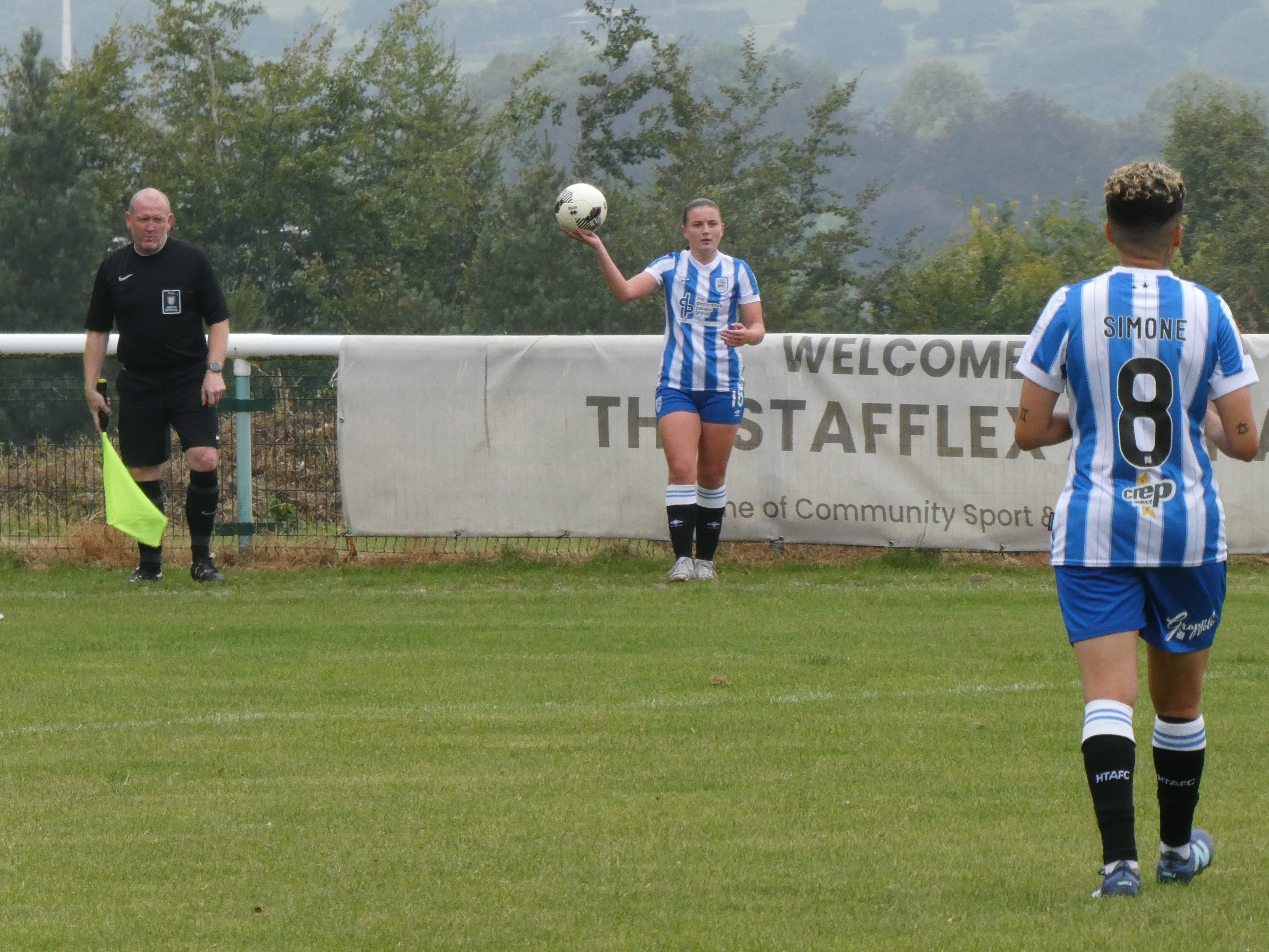 Assistant referee waiting for a footballer to take a throw in with another footballer closer to the camera.