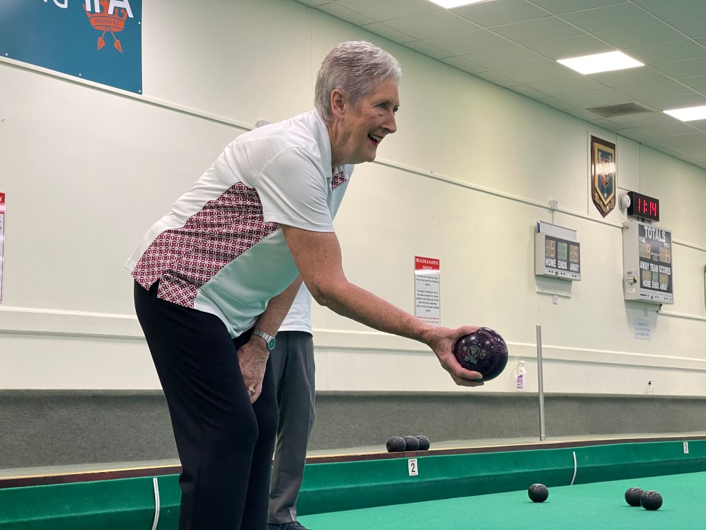 a 73 year old woman holds a bowls ball ready to roll it down to the other end of the green