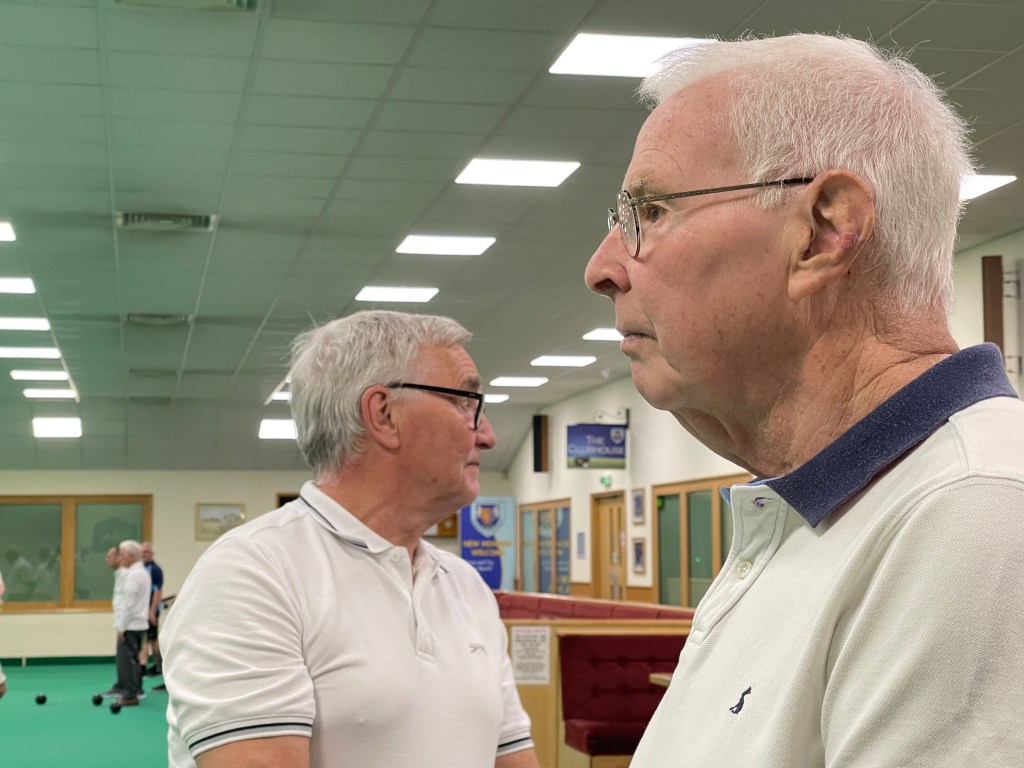 one 80 year old man is having his side profile pictured as he looks on down the green, another man stand behind him looking the other way.